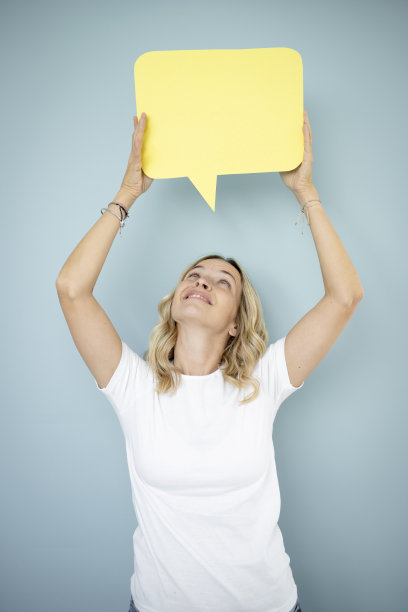 漂亮的金发卷发的年轻女子站在蓝色的墙前，拿着演讲泡沫图片下载