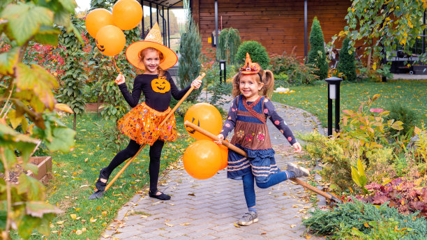 小女巫骑着扫把飞行。万圣节前夕，孩子们打扮成女巫的样子在户外玩耍。快乐的女孩们拿着橙色的气球在花园的小径上欢快地奔跑。图片下载