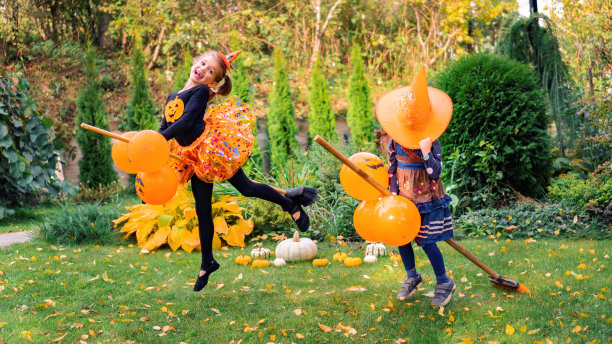 万圣节骑着扫帚飞的小女巫。两个穿着女巫服装的女孩正坐在飞天扫帚上学习飞行。孩子们穿着狂欢节的服装在花园的草坪上扮演女巫。图片下载