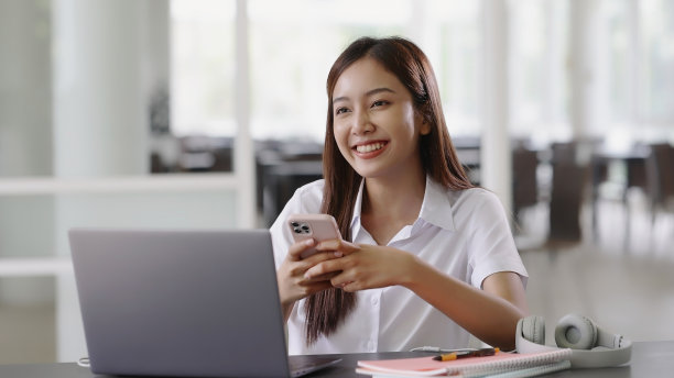 微笑的亚洲美女使用移动智能手机，同时使用笔记本电脑。图片下载