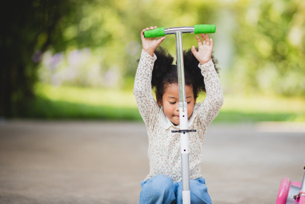 两个俏皮的非洲卷发姐妹骑着滑板车在白天树木环绕的空旷小路上玩耍图片下载