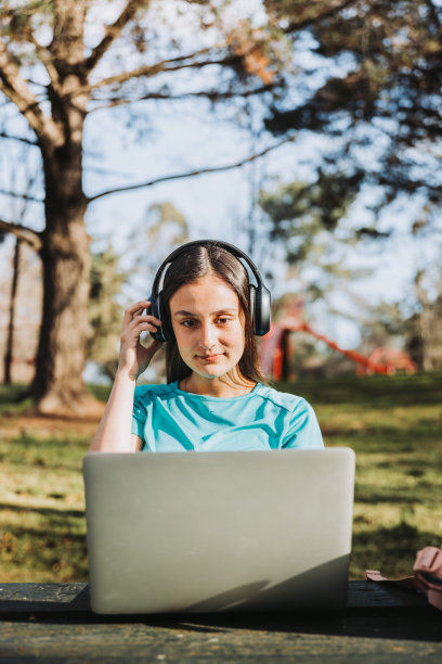 微笑的女大学生在校园自然公园使用笔记本电脑和耳机。电子学习的概念。图片下载