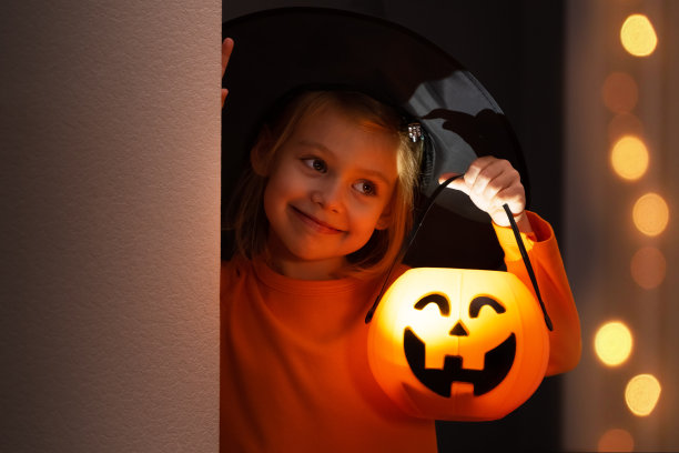 万圣节。快乐的小女孩穿着女巫服，戴着黑帽子，拿着灯笼南瓜，从角落里向外张望，微笑着图片下载
