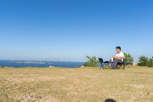 在野外的海边，一名亚洲男子坐在简单的桌椅上喝水图片下载