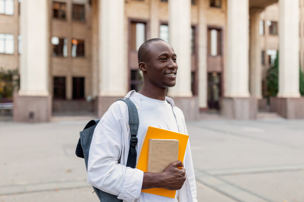 微笑的非裔美国男学生背着背包和工作手册在大学建筑背景摆姿势图片下载