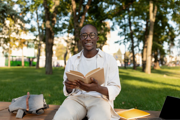 快乐的黑人学生男孩阅读有趣的书，下课后在公园休息，在阳光明媚的日子坐在长凳上图片下载