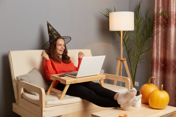 室内拍摄的极为快乐的节日妇女戴着女巫帽在家里的笔记本电脑上工作，握紧拳头，庆祝胜利，在万圣节庆祝期间做她的工作。图片下载