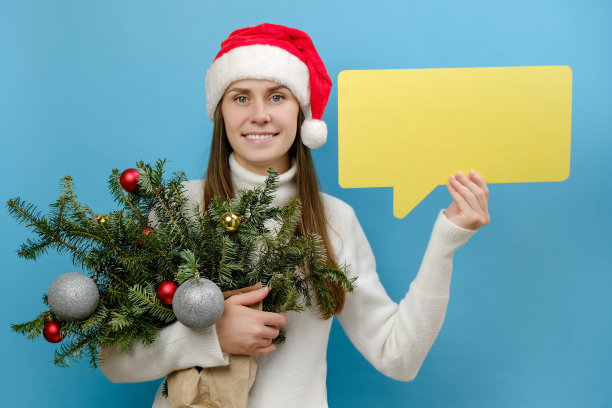 快乐的年轻女孩20多岁在圣诞红帽子拿着一束云杉树枝和黄色的空空白说云说话泡泡，在蓝色的背景。新年喜庆喜庆佳节概念图片下载