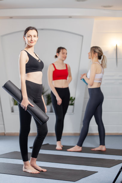 瑜伽教练拿着瑜伽垫摆姿势。背景中有两个女孩在聊天。图片下载