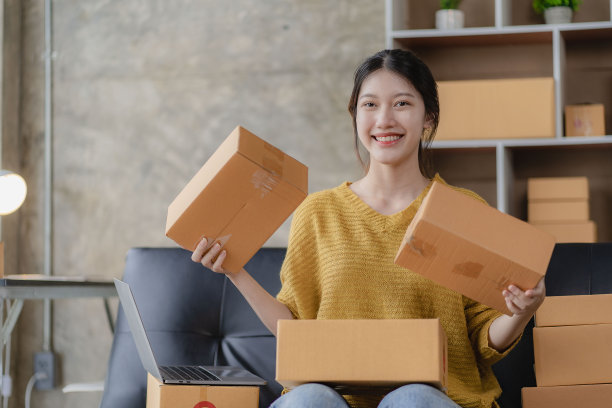 亚洲妇女坐在家里的沙发上自由职业者使用笔记本电脑与盒子，成功的亚洲妇女举手，在线营销包装和送货盒，中小企业的概念图片下载