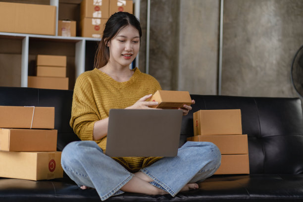 亚洲妇女坐在家里的沙发上自由职业者使用笔记本电脑与盒子，成功的亚洲妇女举手，在线营销包装和送货盒，中小企业的概念图片下载