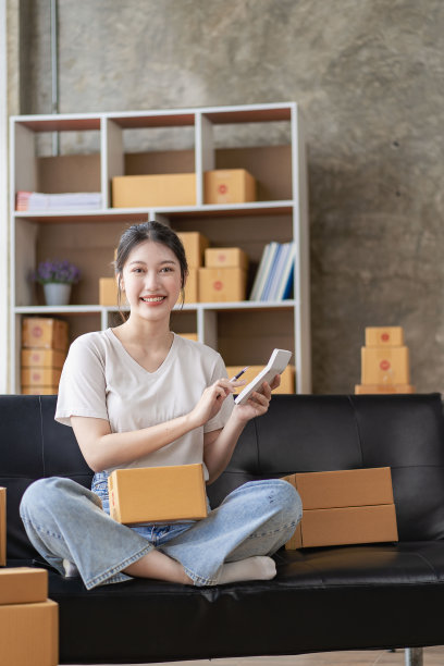 亚洲妇女坐在家里的沙发上自由职业者使用笔记本电脑与盒子，成功的亚洲妇女举手，在线营销包装和送货盒，中小企业的概念图片下载