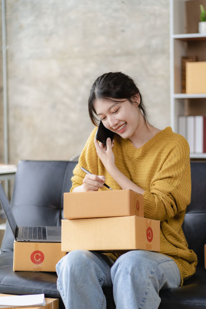 亚洲妇女坐在家里的沙发上自由职业者使用笔记本电脑与盒子，成功的亚洲妇女举手，在线营销包装和送货盒，中小企业的概念图片下载