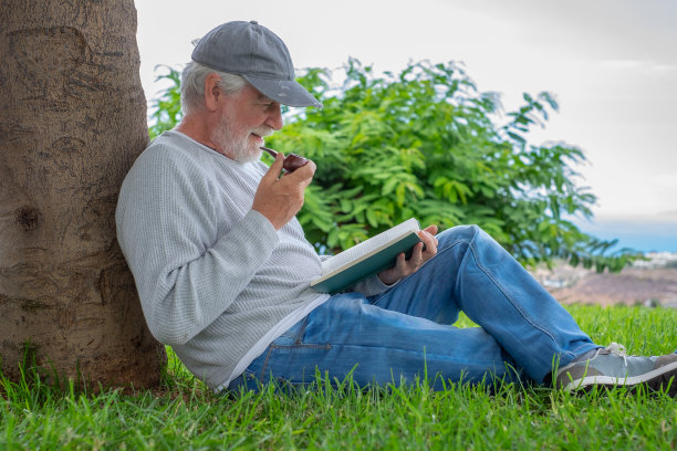 老人的画像，微笑着坐在公园的草地上，悠闲地看书。年长的白种人，有胡子的男性，抽烟斗图片下载