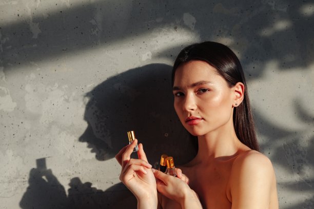 女士手握香水化妆品瓶，用于滴在皮肤或头发上。女人用古龙水在树叶阴影的旧墙背景。健康的生活方式和自我照顾的理念。复制文本空间图片下载