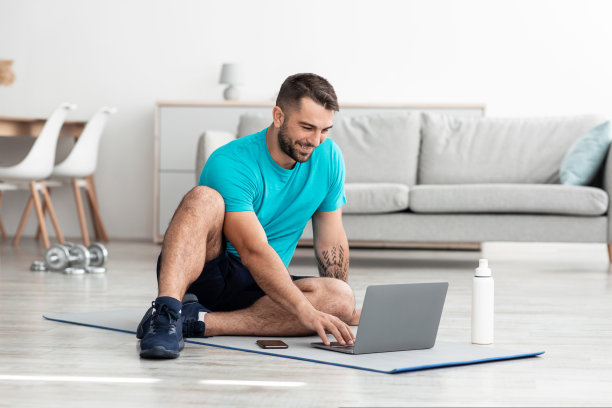 微笑快乐的千禧一代肌肉发达的白人男性博主坐在垫子上，拿着一瓶水和电脑图片下载