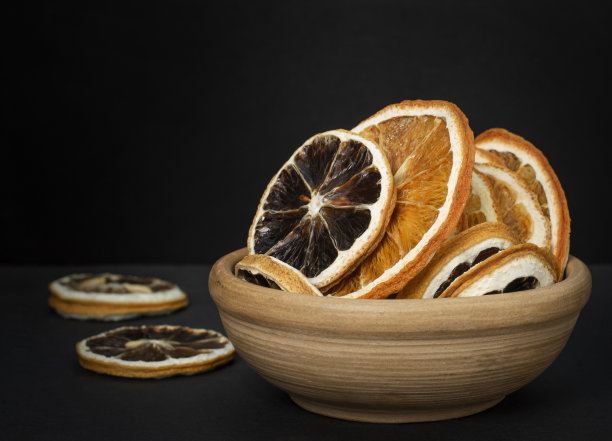 这是一张在深色表面和黑色背景的杯子里的干柠檬和橙子片的特写照片。图片下载