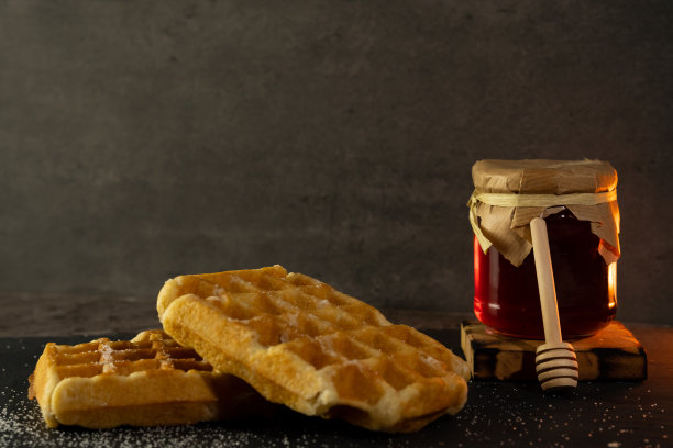 照片静物与新鲜出炉的华夫饼和一罐蜂蜜与蜂蜜棒在黑暗的背景和温暖的光-面包店和糖果的概念图片下载
