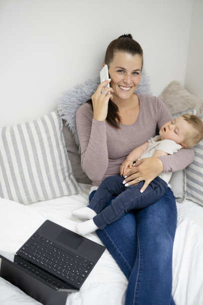 漂亮的年轻母亲坐着，躺在床上，在笔记本上工作，抱着她熟睡的婴儿在她的怀里图片下载