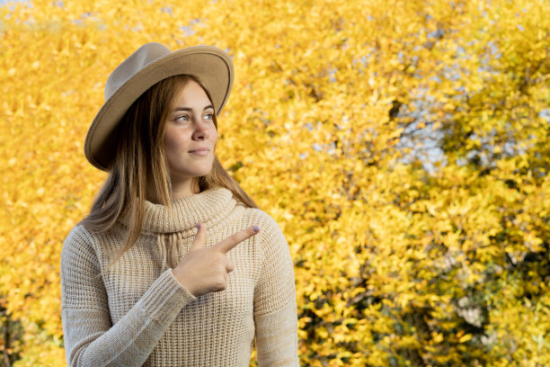 照片可爱迷人的年轻秋天女人在帽子指向空白的黄色树叶背景图片下载