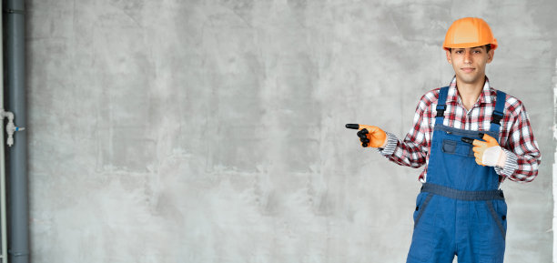 年轻的阿拉伯人越过灰色的墙，穿着承包商制服和安全头盔，开心地微笑着，拇指朝边，开心地笑着。图片下载