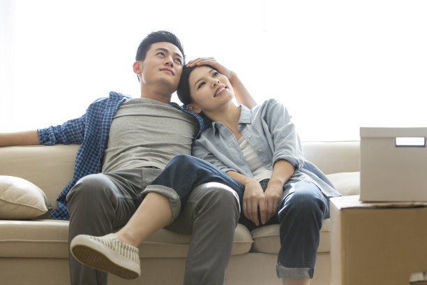 年轻夫妇在新家的沙发上享受生活，妻子依偎在丈夫的怀里，表情充满了渴望和期待——库存照片图片下载