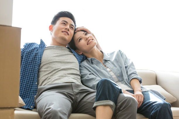 年轻夫妇在新家的沙发上享受生活，妻子依偎在丈夫的怀里，表情充满了渴望和期待——库存照片图片下载