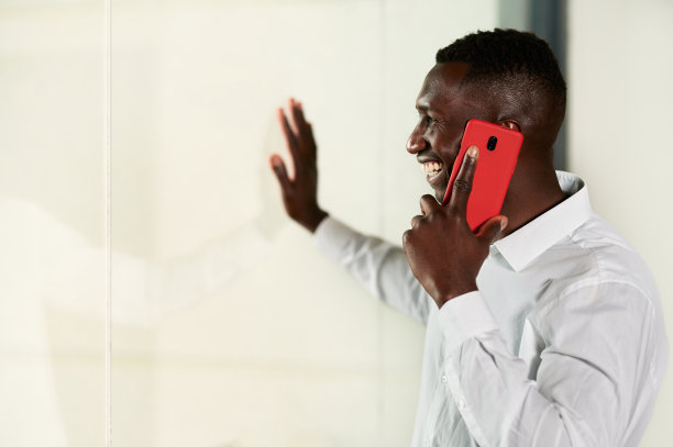 快乐的黑人男子一边用智能手机说话，一边透过大玻璃墙看图片下载