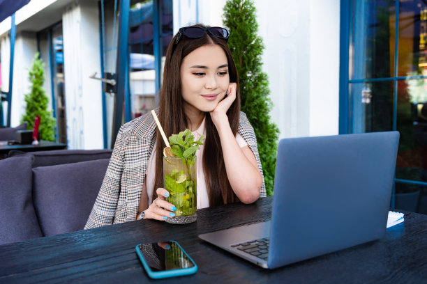 美丽的亚洲女孩拿着一杯莫吉托，看着笔记本电脑上的东西。图片下载