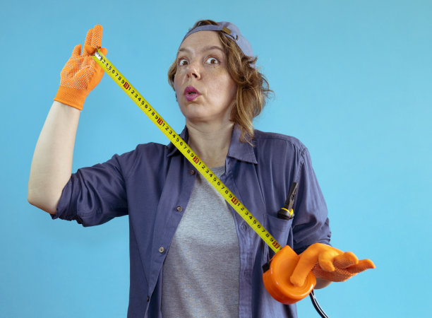 一个成年妇女在她的手中拿着一个橙色手套的建筑卷尺在一个孤立的蓝色背景上。女人的工作。女工头的肖像图片下载