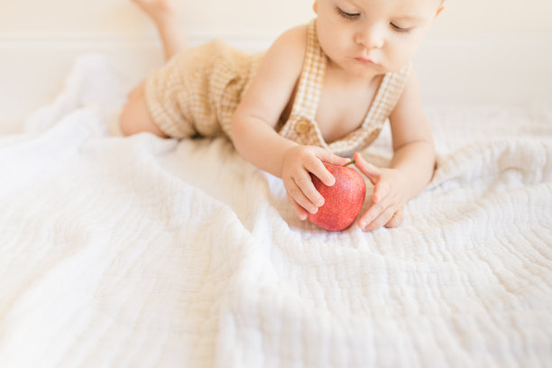 2022年秋季，一个9个月大的流行病男婴穿着黄色格纹工装裤，在家里地板上的奶油色薄纱毯子上玩新鲜的红苹果图片下载