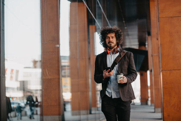 时髦的商人带着皮制公文包在金融区用智能手机听着音频歌曲，戴着耳机喝着咖啡图片下载