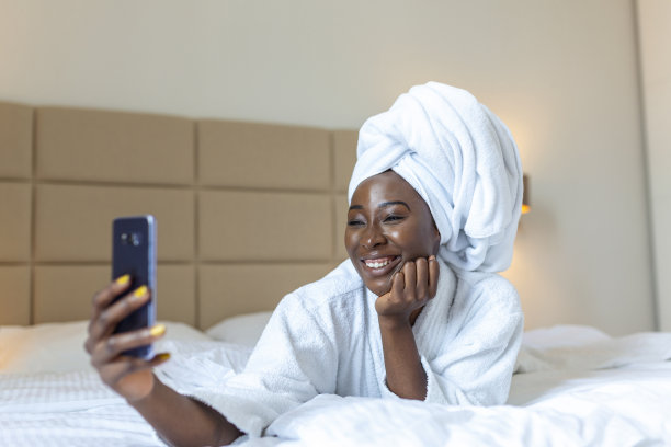 微笑的非洲年轻女子躺在床上，穿着浴袍，用手机自拍。非裔美国妇女沐浴后躺在床上放松，看着手机摄像头自拍图片下载