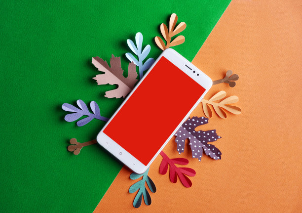 一个白色的手机，中间是红色空白的文字，放在半绿色和橙色的背景上，周围是红色、绿色、蓝色和棕色的纸叶。秋天的概念图片下载