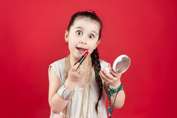 黑头发的小女孩在嘴唇上涂了红色的口红。有趣的照片拍摄在工作室的红色背景图片下载