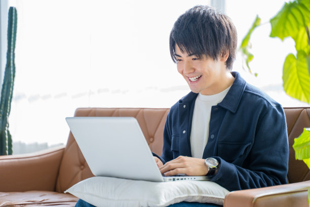 一个20多岁的男人一边在客厅沙发上用笔记本电脑工作，一边给植物看叶子图片下载