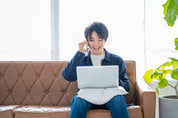 一个20多岁的男人一边在客厅沙发上用笔记本电脑工作，一边给植物看叶子图片下载