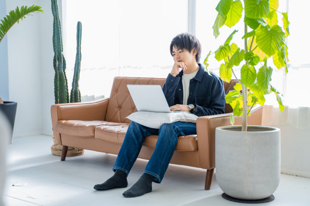 一个20多岁的男人一边在客厅沙发上用笔记本电脑工作，一边给植物看叶子图片下载