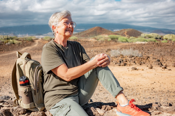 微笑的老妇人在户外远足坐在一堵石墙看向别处。活跃成熟的女人坐在她的背包附近享受自由和阳光灿烂的日子图片下载