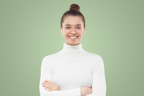 年轻女子穿着白色高领毛衣，扎着头，双臂交叉站着，面带自信的微笑图片下载