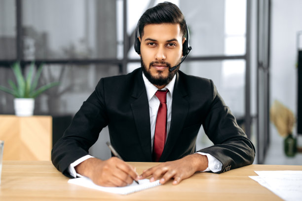 成功的严肃的印度或阿拉伯年轻商人的照片，金融导师，戴着耳机，坐在他现代办公室的桌面，看着相机，听着对话者，做着笔记图片下载