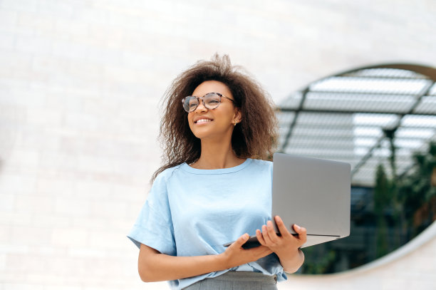 照片可爱优雅的非裔美国年轻女子卷头发，在蓝色衬衫和眼镜，站在户外，拿着一个打开的笔记本电脑，看向一边，快乐地微笑，梦想，思考图片下载