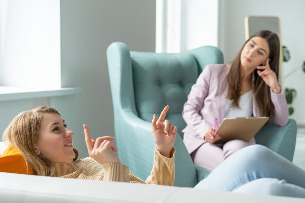 穿着米色毛衣和蓝色牛仔裤的快乐女病人，一边打着手势，一边躺在沙发上谈论她的问题，这是心理医生的预约。心理学家坐在对面的椅子上。图片下载