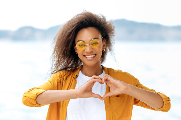 照片上是一个美丽、积极、时尚的卷发非洲裔美国女孩，戴着橙色眼镜，站在户外，用她的手画出一个心形，这是爱、和平的象征，她看着镜头，微笑着图片下载