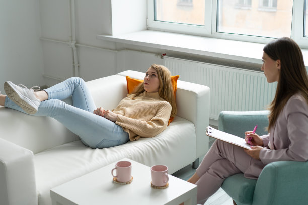 一个穿着蓝色牛仔裤和芥末色运动衫的女人躺在心理医生预约的沙发上。谈论他的问题。穿着粉红色西装的心理学家坐在对面的椅子上。明亮宽敞的办公室。模糊的前景。图片下载