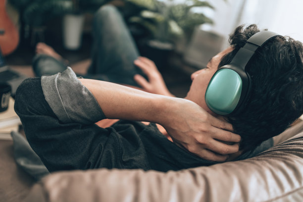 男人戴着耳机听着音乐，躺在家里的沙发上脖子疼。放松的家的概念。图片下载