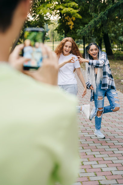 一个男孩的手拿着手机，在公园里给他的两个疯狂的女朋友拍照。暑假的乐趣。移动电话通信。的乐趣。快乐女生朋友在一起。图片下载