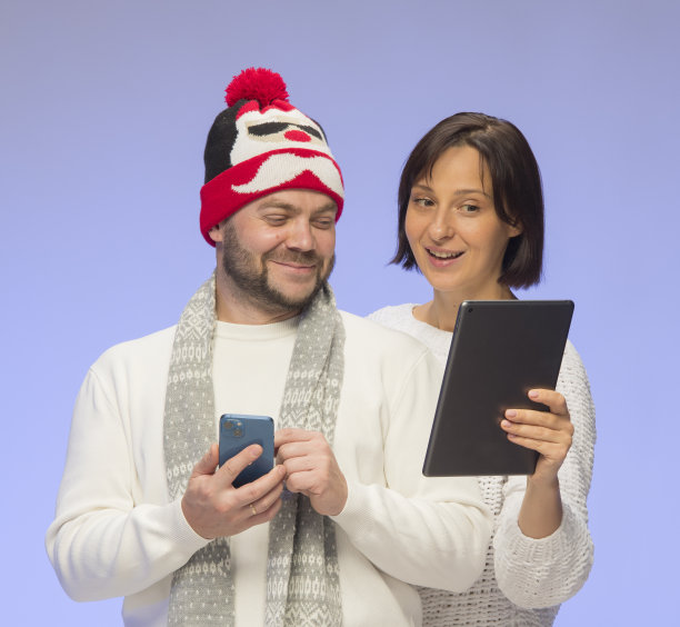 快乐的微笑的夫妇男人和女人礼品盒和智能手机平板电脑在蓝色背景的白色毛衣孤立。网上购物圣诞礼物概念图片下载