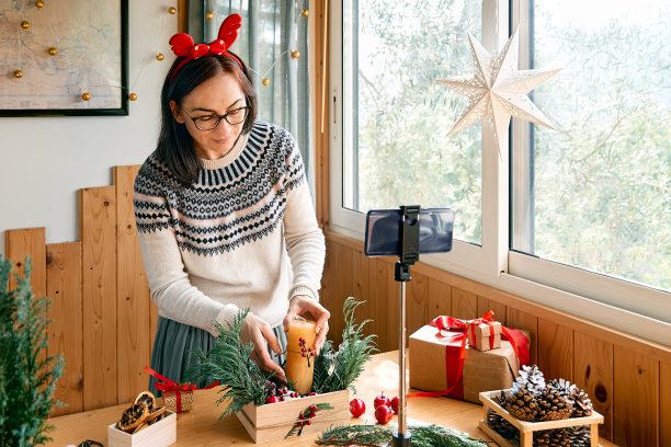 女花匠博主在她的智能手机前用新鲜的松枝、蜡烛和圣诞装饰制作冬季花道。小生意。季节性冬季车间。图片下载