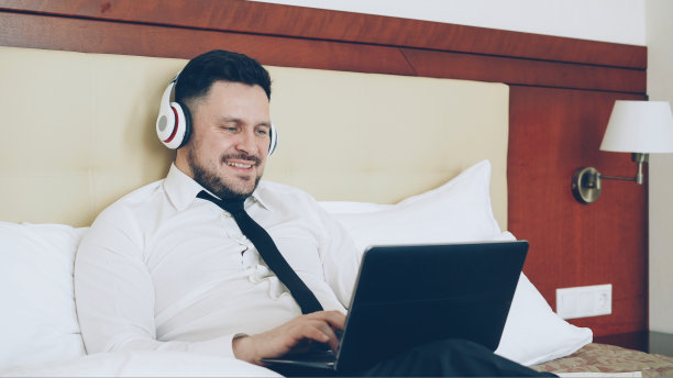 快乐的商人戴着耳机在笔记本电脑前工作，听着音乐，躺在酒店房间的床上微笑。旅游，商务和人的概念图片下载